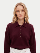 A woman with blonde hair wearing a dark burgundy knit polo shirt with brown buttons and white pants, photographed against a light gray studio background.