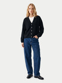 A woman stands against a light gray studio background wearing a black button-front cardigan over a white top, blue jeans, and black shoes.