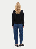 A woman viewed from behind wearing a black long-sleeve top, blue jeans, and black loafers against a light gray studio background.
