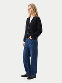 A woman stands in profile wearing a black button-front cardigan over a white top, blue wide-leg jeans, and black shoes, photographed against a plain light gray studio background.
