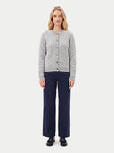 A woman stands against a light gray studio background wearing a light gray cable-knit button-front cardigan, dark navy wide-leg trousers, and black shoes.