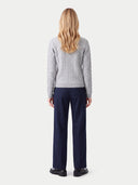 A woman viewed from the back wearing a light gray cable-knit sweater and navy blue trousers, standing against a plain light gray studio background.