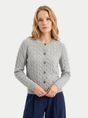 A woman wearing a light gray cable-knit button-up cardigan sweater and navy blue trousers, standing against a plain light gray studio background.