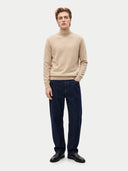 A male model wearing a beige ribbed turtleneck sweater, dark navy blue jeans, and black leather shoes, standing against a light gray studio background.
