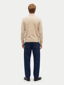 A man viewed from the back wearing a beige mock-neck knit sweater and dark blue jeans, standing against a light gray studio background.