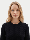 A blonde woman wearing a black cable-knit crewneck sweater, photographed against a clean, light gray studio background.