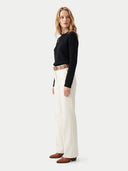 A woman modeling a black ribbed long-sleeve top and off-white wide-leg trousers with a brown belt, standing against a light gray studio background.