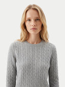 A blonde woman wearing a light gray cable-knit crewneck sweater, shown from the chest up against a plain light gray studio background.