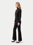 A woman in a black mock-neck knit sweater and black flared trousers, standing in profile against a light gray studio background.