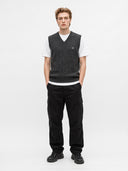 A male model stands against a light gray studio background wearing a dark gray textured knit sweater vest over a white t-shirt, paired with black cargo pants and black sneakers.