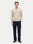 A male model wearing a cream cable-knit mock-neck sweater with a three-button placket, paired with navy trousers and black shoes, standing against a light gray studio background.