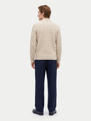A man viewed from the back wearing a cream cable-knit mock-neck sweater and navy blue trousers, standing against a light gray studio background.