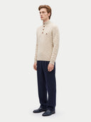 A man wearing a cream cable-knit mock-neck sweater with brown buttons, navy trousers, and black leather loafers, standing against a light gray studio background.