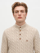 A man wearing a beige cable-knit mock-neck sweater with a three-button placket and a small embroidered logo on the chest, photographed against a plain light gray background.