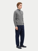 A man wearing a gray cable-knit mock-neck sweater and navy blue trousers, standing in profile against a light gray studio background.