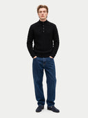 front view of a man standing wearing a black cable knit quarter buttoned cardigan and blue jeans with navy flats on a light gray background.