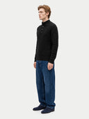 A man wearing a black cable-knit mock-neck sweater, blue jeans, and dark blue slip-on shoes, standing in profile against a light gray studio background.