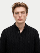 A young man with light brown hair wearing a black cable-knit polo shirt, photographed against a solid light gray background.