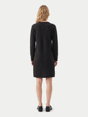 A woman viewed from the back wearing a dark charcoal knit long-sleeve dress and black loafers, standing against a light gray studio background.