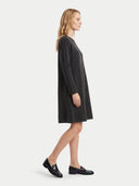 A woman in profile wearing a dark charcoal gray long-sleeve shift dress and black leather loafers, standing against a light gray studio background.