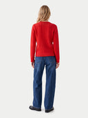 A woman viewed from the back wearing a bright red cable-knit sweater and blue jeans against a light gray studio background.
