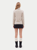 A woman viewed from the back wearing a light beige turtleneck sweater, a black mini skirt, white ribbed knee-high socks, and black ankle boots, against a plain light gray studio background.
