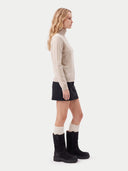 A woman in side profile wearing a beige turtleneck sweater, a black mini skirt, cream ribbed socks, and black boots, photographed against a plain light gray studio background.