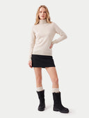A woman modeling a light beige ribbed turtleneck sweater, paired with a black mini skirt, cream ribbed socks, and black ankle boots, against a plain light gray studio background.