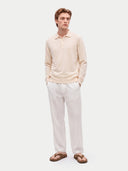 A man modeling a light beige long-sleeve polo sweater and off-white trousers, paired with brown sandals, against a solid light gray background.