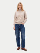 A woman modeling a beige mock-neck knit sweater and blue jeans with brown leather shoes, standing against a light gray studio background.