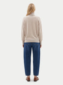 A woman viewed from the back wearing a light beige turtleneck sweater, blue jeans, and brown loafers against a plain light gray studio background.