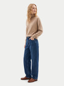 A woman stands in profile wearing a beige ribbed turtleneck sweater, wide-leg blue jeans, and brown leather loafers against a plain light gray studio background.