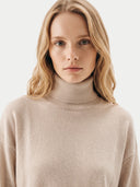A close-up shot of a woman with blonde hair wearing a light beige ribbed turtleneck sweater against a plain white background.