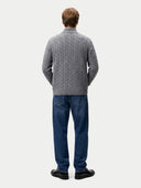 A man viewed from behind wearing a gray cable-knit sweater, blue jeans, and black shoes against a light gray studio background.
