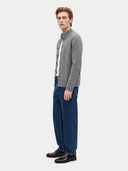 A man modeling a gray cable-knit cardigan over a white shirt, paired with dark blue jeans and black leather shoes, against a light gray studio background.