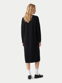 A woman with blonde hair is shown from the back wearing a long-sleeved black midi dress, white socks, and black loafers against a light gray studio background.