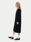 A woman in profile wearing a long black dress, white socks, and black loafers, standing against a plain light gray studio background.