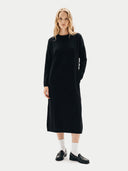 A blonde model wearing a long-sleeve black midi dress with white socks and black loafers, standing against a light gray studio background.