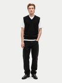 A male model stands against a light gray studio background wearing a black knit sweater vest over a white short-sleeve crewneck t-shirt, paired with black trousers and black leather dress shoes.