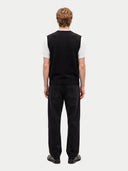 back view of a man standing wearing a black vest over a white t-shirt, black jeans, boots on a light gray background.