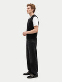 A man stands in profile wearing a black knit sweater vest over a white short-sleeve t-shirt, black wide-leg jeans, and black leather loafers, against a light gray studio background.