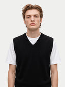 A young man wearing a black V-neck knit sweater vest over a white crew-neck t-shirt, photographed against a solid light gray background.