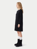 A woman stands in profile wearing a black long-sleeve knit dress with a relaxed, knee-length silhouette, paired with white socks and black ankle boots, against a plain light gray studio background.
