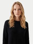 A blonde woman with wavy hair wearing a black crewneck sweater, photographed against a clean light gray background.