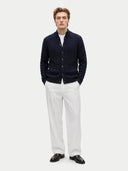 A man modeling a dark navy blue cable-knit button-up cardigan over a white t-shirt, paired with white trousers and black leather loafers, against a light gray studio background.