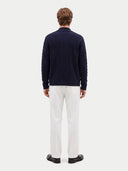 A man viewed from behind wearing a navy blue cable-knit sweater, white trousers, and black shoes against a light gray studio background.