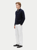A man wearing a dark navy blue textured knit cardigan over a white collared shirt, paired with white trousers and black leather loafers, standing against a light gray studio background.