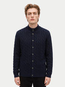 A man wearing a navy blue cable-knit button-up cardigan sweater with a mock neck, photographed against a light gray studio background.
