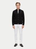 A man modeling a black cable-knit button-up cardigan sweater, styled with white trousers and black leather loafers, against a light gray studio background.