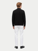 A man viewed from the back wearing a black cable-knit sweater, white trousers, and black shoes against a light gray studio background.